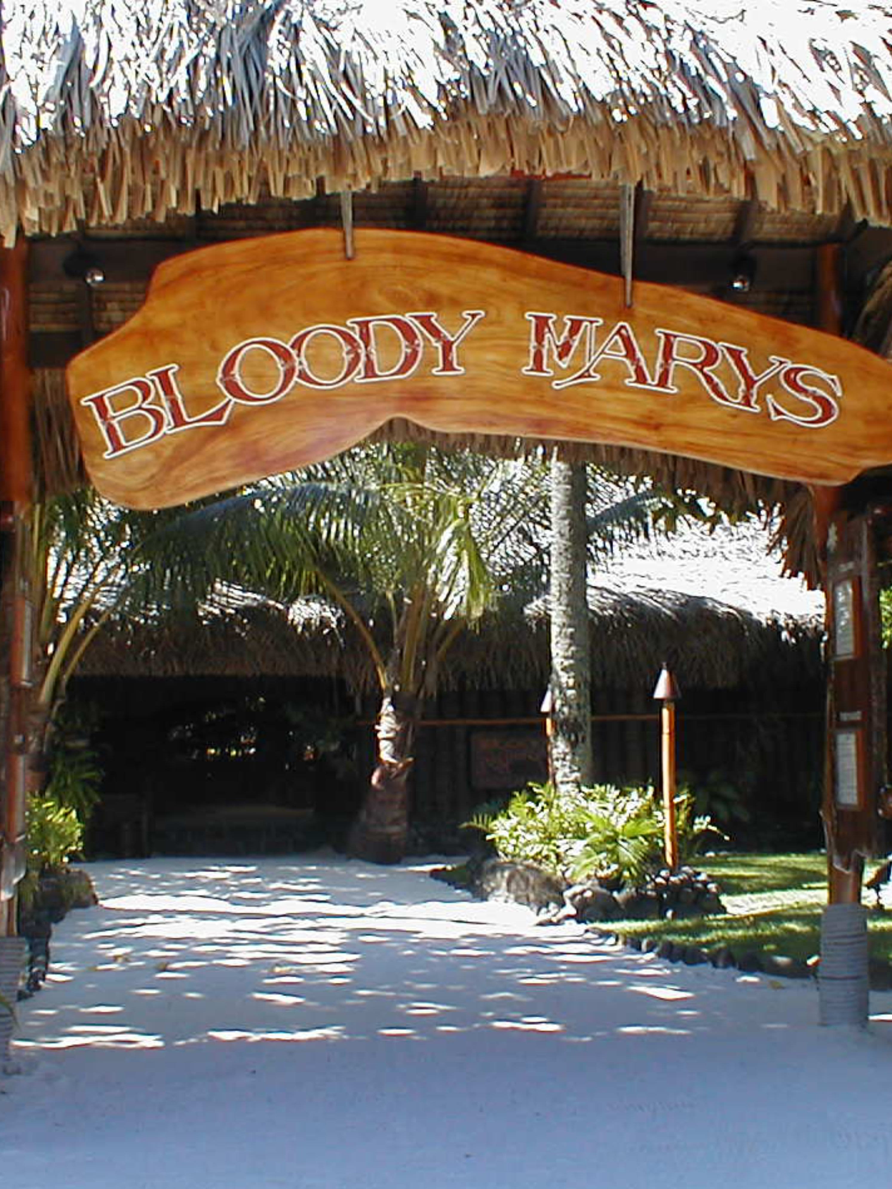 Entrée du Bloody Mary's avec grande enseigne en bois, toit en chaume, chemin de sable et cocotiers