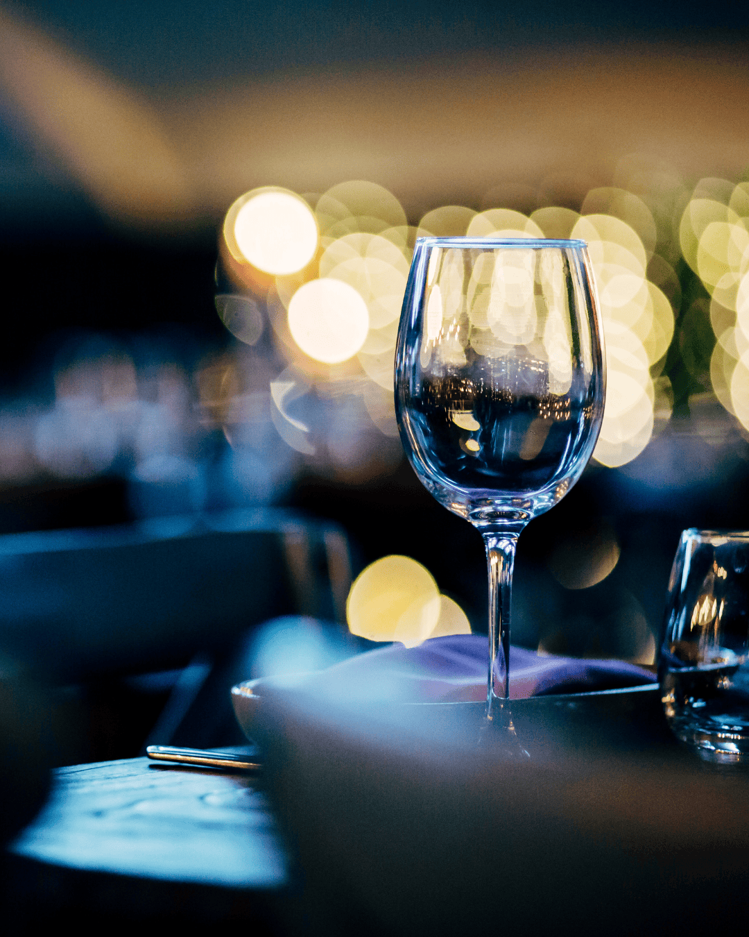 Verre à pied sur une table dressée dans une ambiance de soirée, lumières dorées floues en arrière-plan