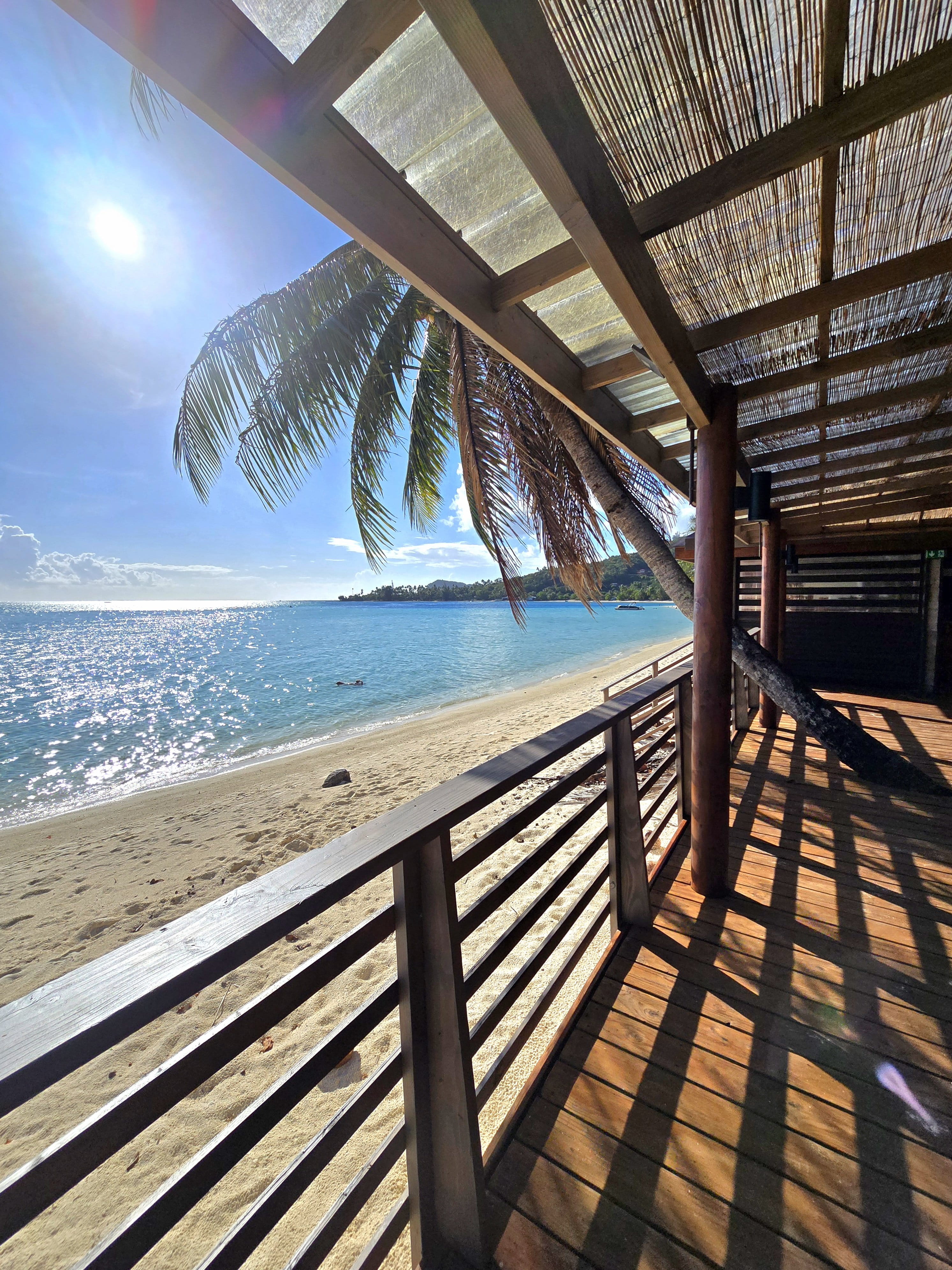 Vue depuis une terrasse bois ombragée vers la plage de sable fin, cocotiers et lagon turquoise (La Bella Matira)