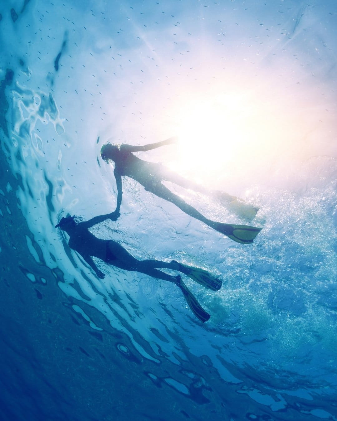 Vue sous la surface — silhouettes mains jointes de deux personnes en masque et tuba contre une eau turquoise striée par des rais lumineux.