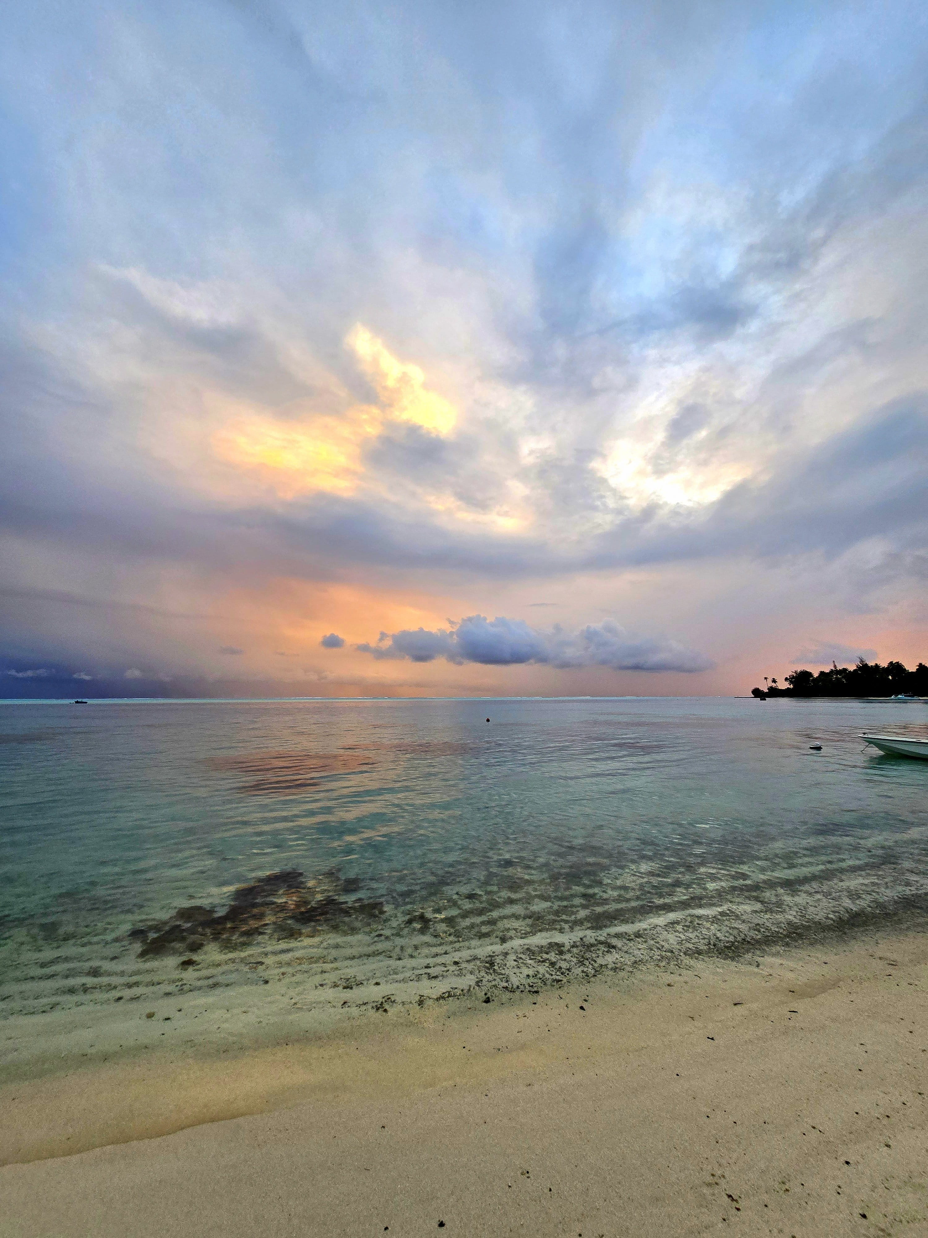 Bord de lagon paisible se fondant dans du sable pâle sous des couches de nuages pastels dorés ou rosés comme au lever ou au coucher du soleil, lointaine presqu’île boisée et bateaux sur le récif.
