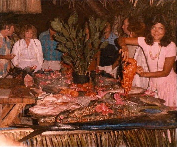 Scène retro d’un banquet marin sur glace sous un toit de chaume, étal garni de fleurs dont hibiscus, une femme brandit fièrement une langouste parmi les convives en tenues lumineuses, tous très souriants.
