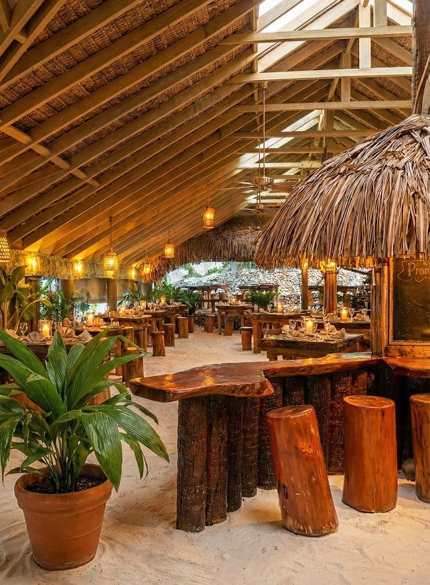 Intérieur de restaurant tout en atmosphère au sol sablonneux avec comptoir en bois sculpté, tabourets en rondins, plafond à pointes de chaume, lanternes suspendues en fibres tressées et chandelles parmi verdure tropicale.