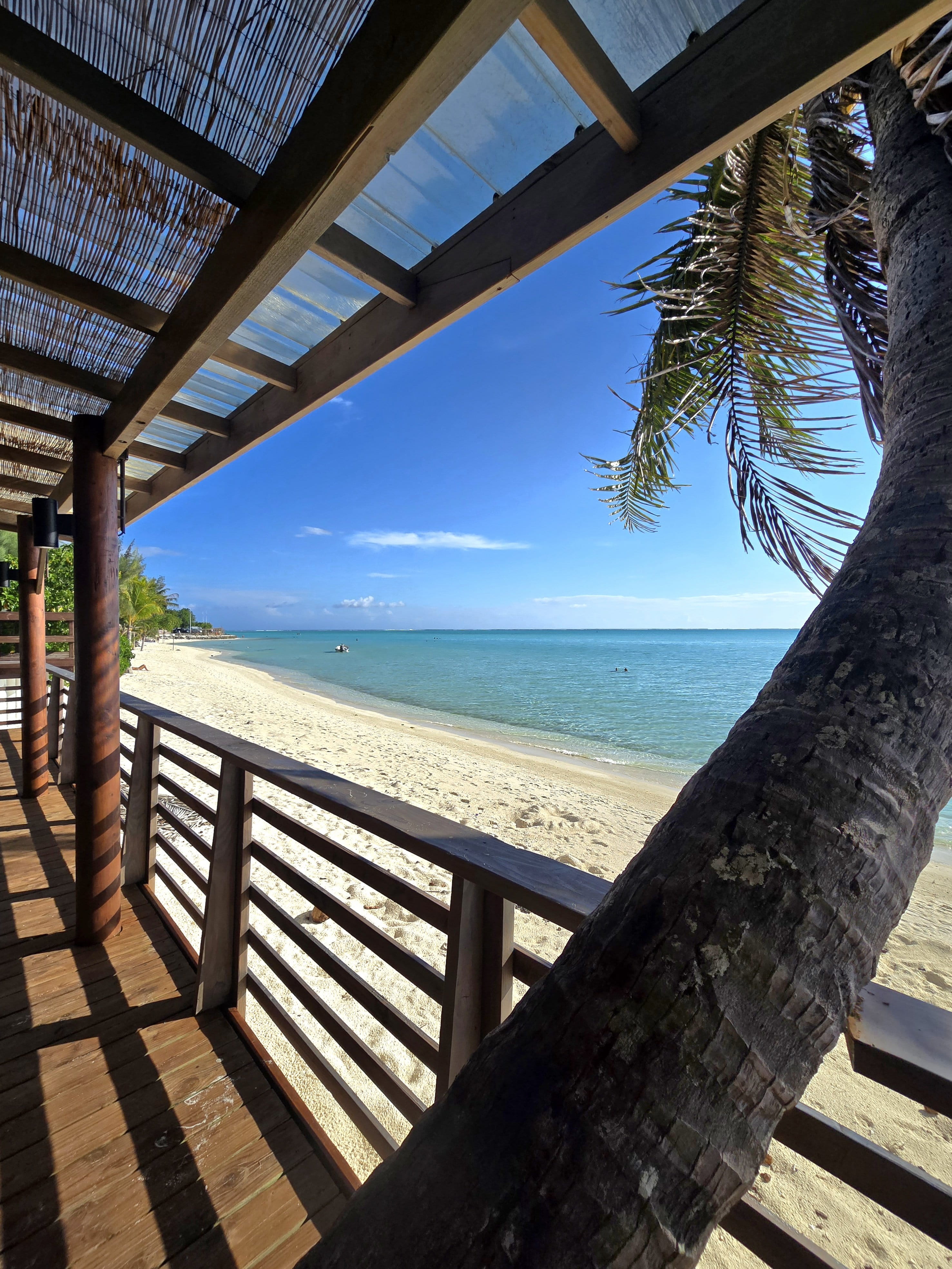 Grande terrasse en bois ombrée, palmier tout près, plage blonde et lagon turquoise jusqu’aux petites barques au loin.