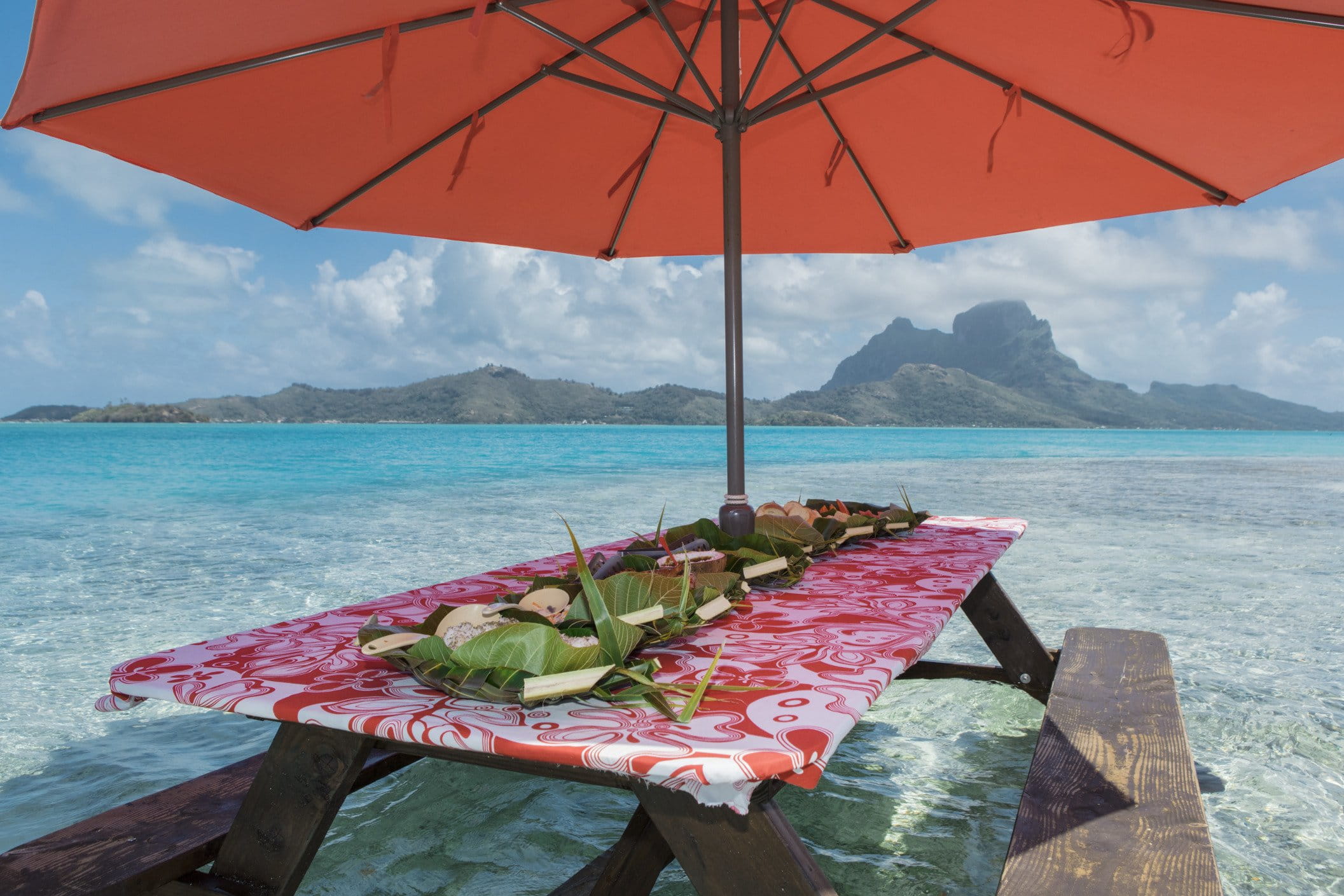 Pause gourmande dans le lagon peu profond — parapluie orange, table quasi immergée, plateaux façonnés en feuilles de bananier et fruits frais avec relief volcanique à l’horizon.