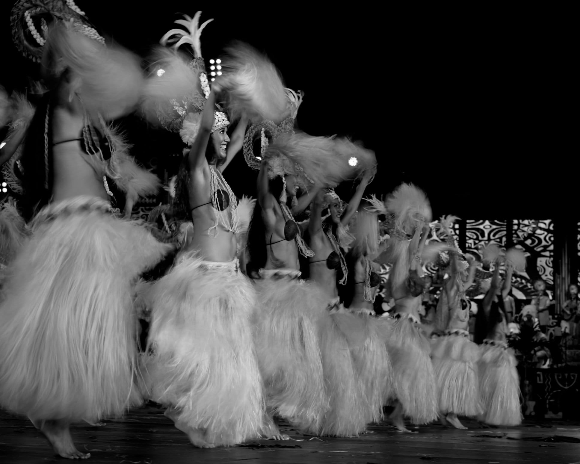 Photo en noir et blanc de danseuses tahitiennes en costume traditionnel sur une scène de nuit.