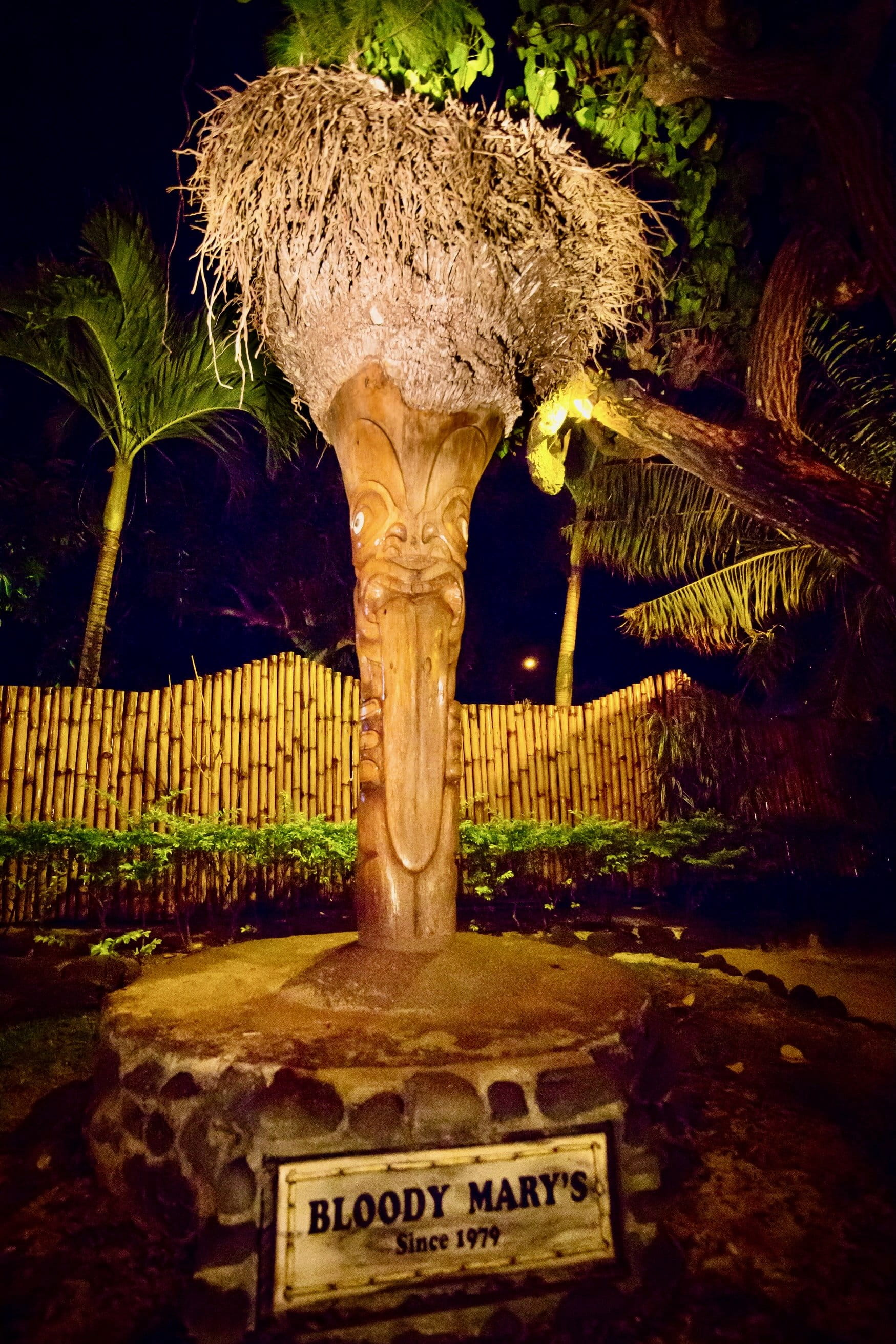 Tiki en bois sculpté de nuit avec enseigne Bloody Mary's et Since 1979 sur le socle en pierre.