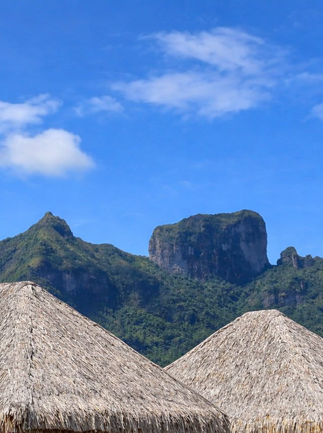 Toits de fare polynésiens en chaume avec le mont Otemanu et les crêtes boisées sous un ciel bleu.