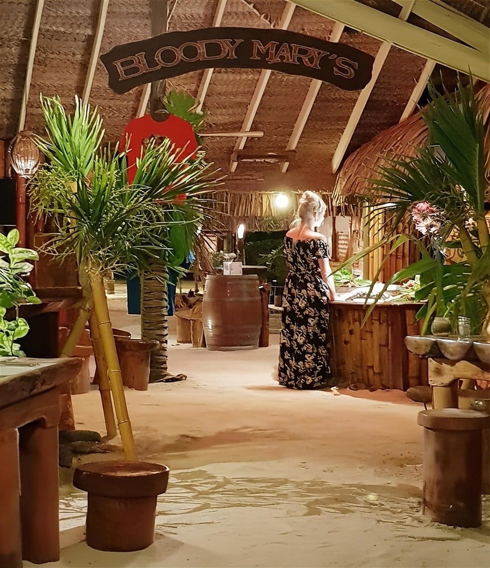 Intérieur du restaurant Bloody Mary's avec sol en sable, client au bar en bois et enseigne sculptée.