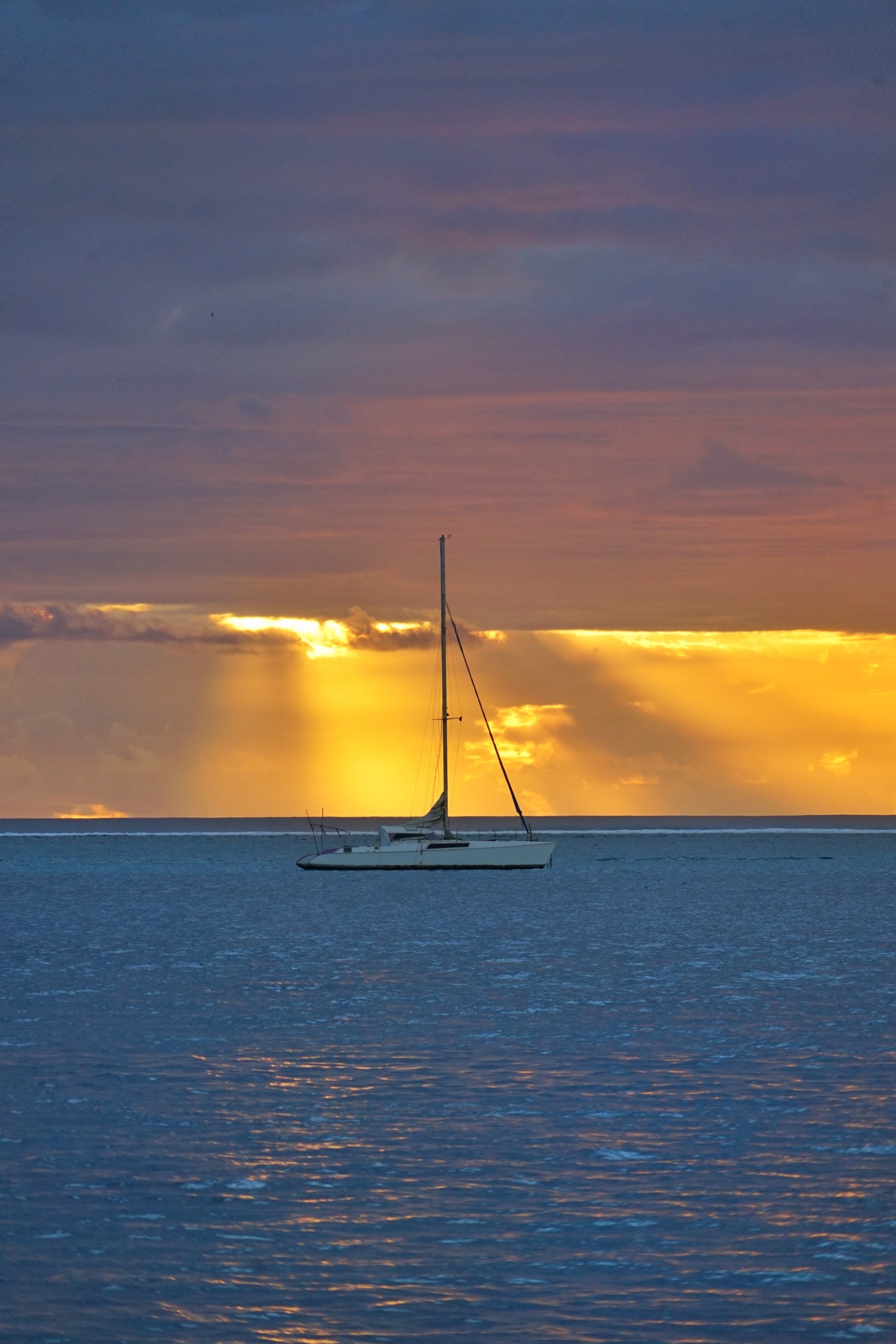 Catamaran blanc sur une mer calme au coucher du soleil, rayons dorés à travers des nuages mauves.