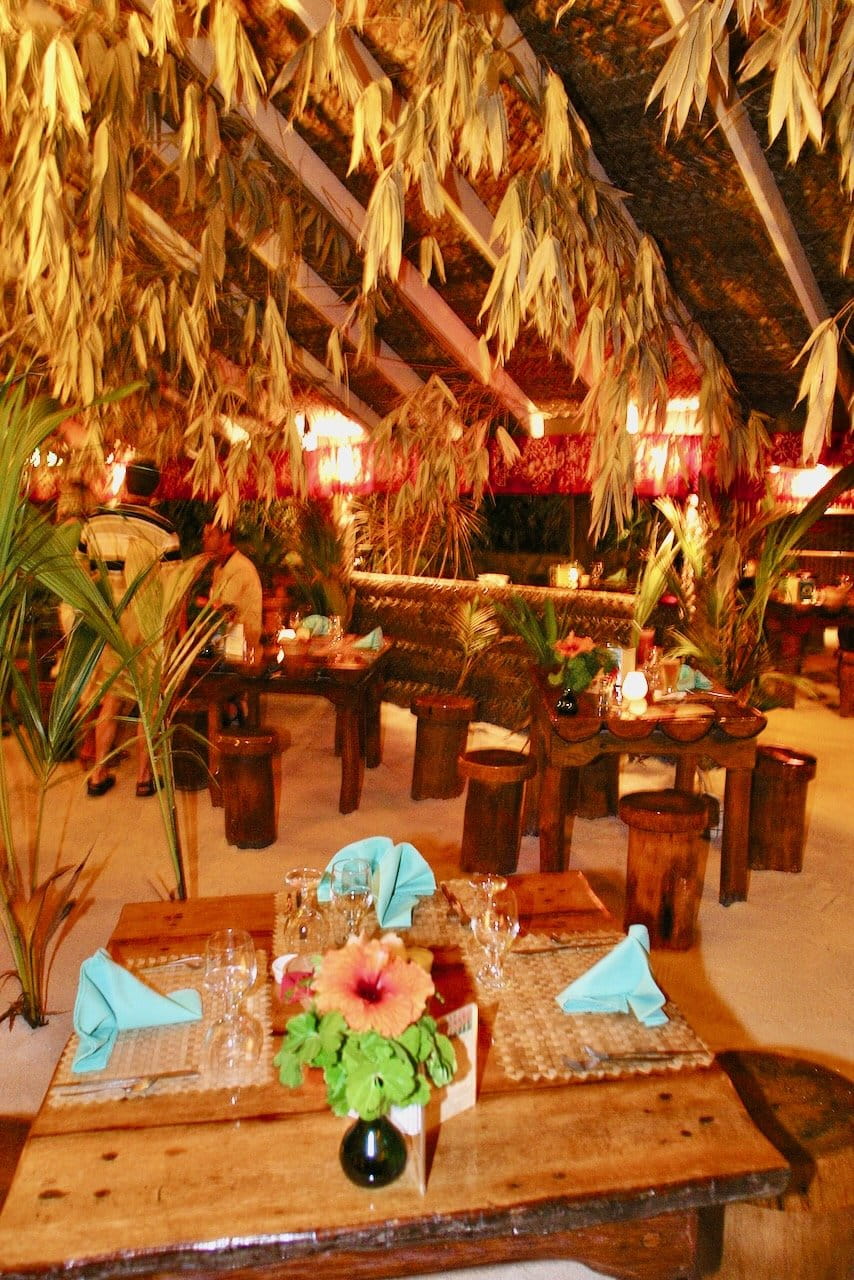 Candle-lit sand-floor dining room with massive sculpted tables beneath vaulting pandanus roofs