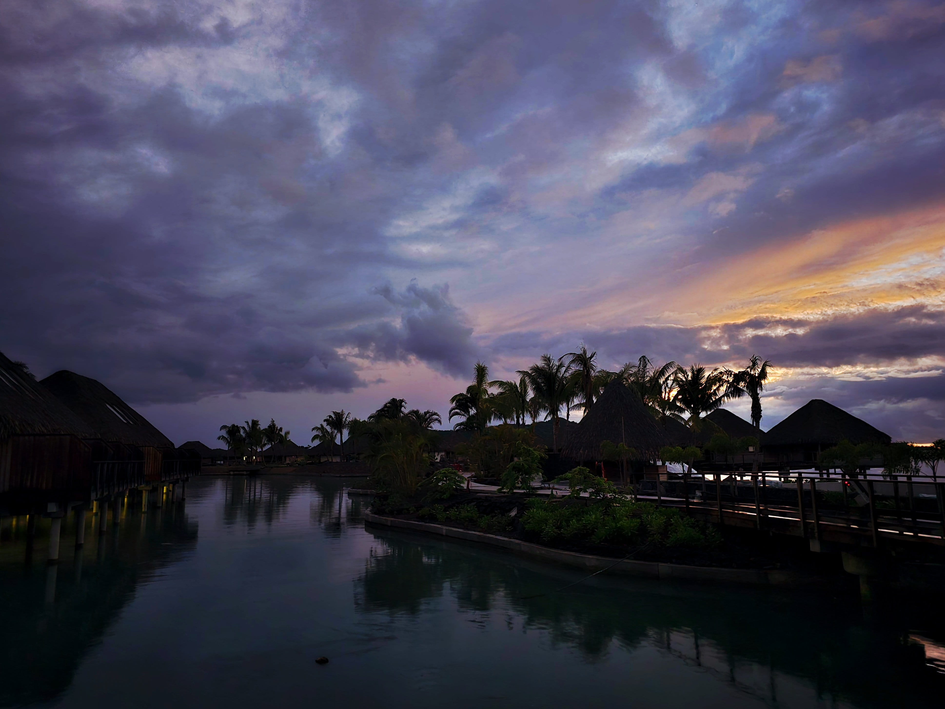 Crépuscule sur des bungalows sur pilotis en silhouette, lagon calme, ciel violet et doré.