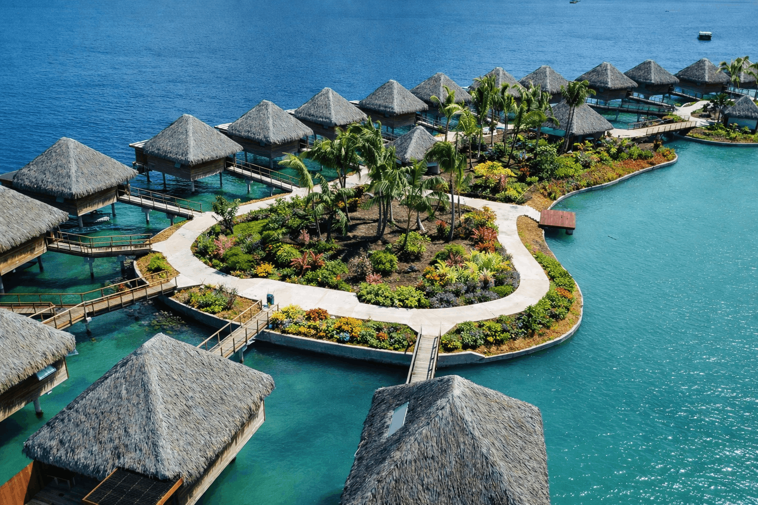 Vue aérienne de bungalows sur pilotis reliés par des passerelles autour d'un îlot paysagé dans une eau turquoise.